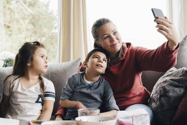 Tochter sieht Mutter an, die ein Selfie mit ihrem autistischen Bruder macht, während sie bei ihrem Vater im Wohnzimmer sitzt - MASF13907