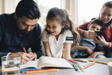Vater bringt seiner Tochter das Zeichnen bei, während die Frau mit ihrem autistischen Sohn im Hintergrund ein Smartphone benutzt - MASF13908
