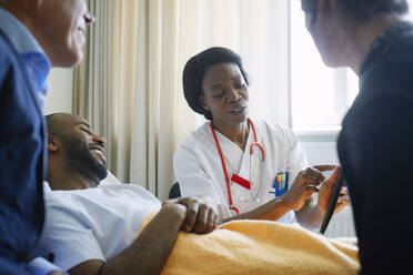 Female doctor explaining medical record over digital tablet to patient and family in hospital ward - MASF13968