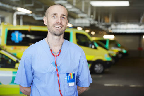 Portrait of smiling male doctor with stethoscope against ambulance in parking lot - MASF13993