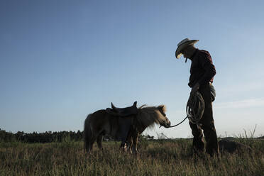 Cowboy mit gesatteltem Pony in einem sonnigen ländlichen Feld - FSIF04431