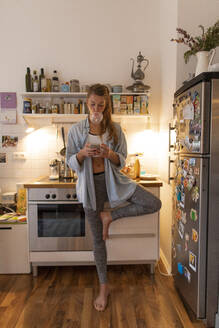 Junge Frau in Yogapose prüft Handy in der Küche zu Hause - GUSF02460