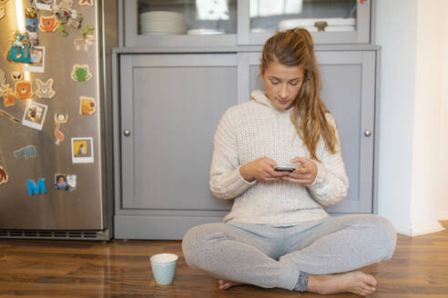 Junge Frau sitzt auf dem Boden in der Küche zu Hause und benutzt ein Mobiltelefon - GUSF02474