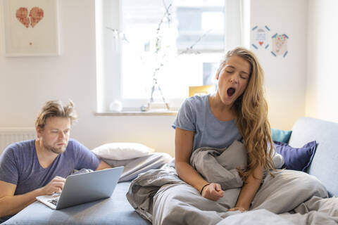 Junge Frau zu Hause, die mit ihrem Partner aufwacht und einen Laptop benutzt, lizenzfreies Stockfoto