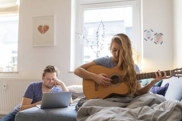 Young woman at home playing guitar with damaged string with partner using laptop - GUSF02486