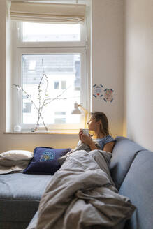 Young woman lying on couch at home looking out of window - GUSF02488