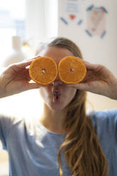 Playful young woman covering her eyes with oranges at home - GUSF02500