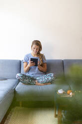 Smiling young woman sitting on couch at home using tablet - GUSF02504