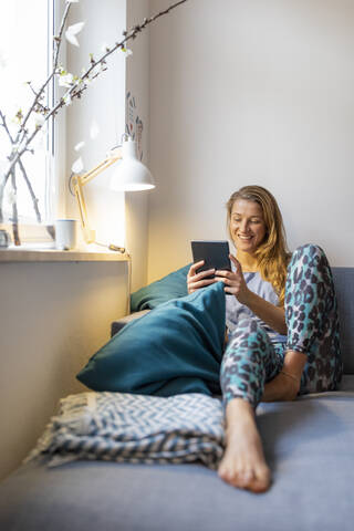 Glückliche junge Frau sitzt zu Hause auf der Couch und benutzt ein Tablet, lizenzfreies Stockfoto