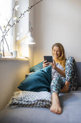 Happy young woman sitting on couch at home using tablet - GUSF02506