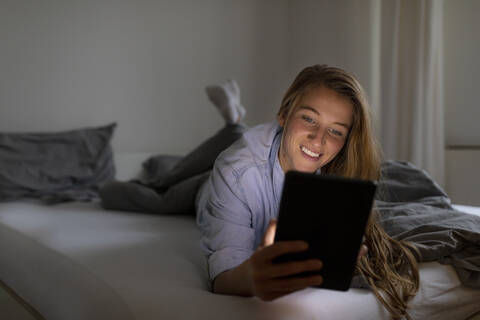 Glückliche junge Frau, die nachts zu Hause im Bett liegt und ein Tablet benutzt, lizenzfreies Stockfoto