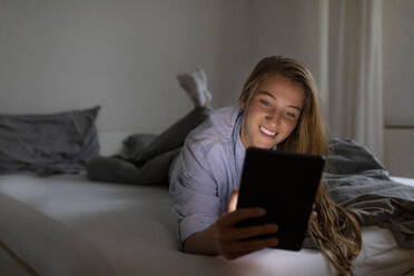 Happy young woman lying in bed at home at night using tablet - GUSF02510