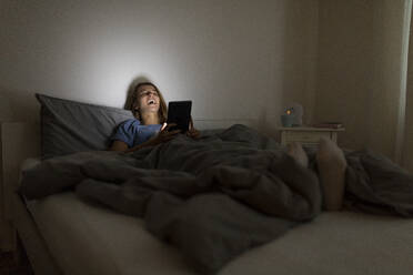 Laughing young woman lying in bed at home at night using tablet - GUSF02514