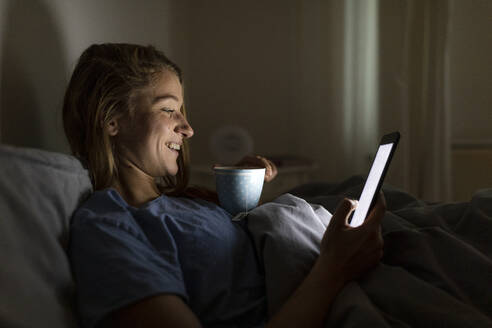 Young woman lying in bed at home with cup of tea at night using tablet - GUSF02515