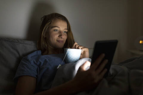 Junge Frau zu Hause im Bett liegend mit einer Tasse Tee und einem Tablet, lizenzfreies Stockfoto