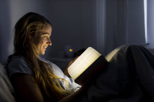 Lächelnde junge Frau, die zu Hause im Bett ein beleuchtetes Buch liest - GUSF02528