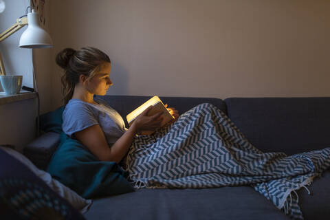Junge Frau liest illuminiertes Buch auf der Couch zu Hause, lizenzfreies Stockfoto