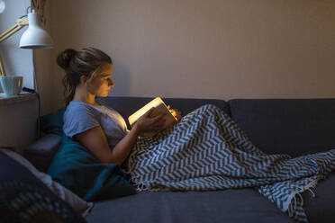 Junge Frau liest illuminiertes Buch auf der Couch zu Hause - GUSF02531