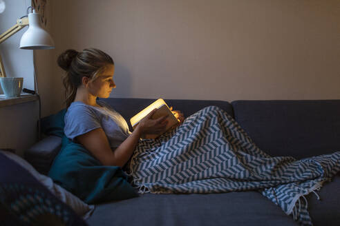 Young woman reading illuminated book on couch at home - GUSF02531