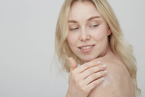 Portrait of blond young woman applying cream on her shoulder - PGCF00023