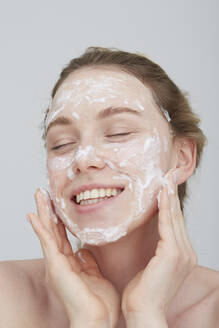 Portrait of blond young woman with eyes closed applying cream on her face - PGCF00028