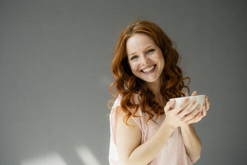 Portrait of happy redheaded woman with cereal bowl against grey background - KNSF06491