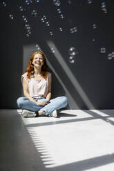 Portrait of happy woman sitting on the floor with coffee bowl watching soap bubbles - KNSF06538