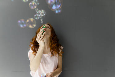 Redheaded woman leaning against grey wall blowing soap bubbles - KNSF06545