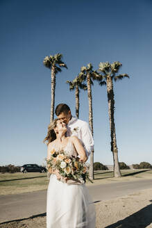 Glückliche Braut und Bräutigam umarmen sich an Palmen unter blauem Himmel - LHPF00789
