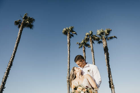 Glückliche Braut und Bräutigam umarmen sich an Palmen unter blauem Himmel - LHPF00790