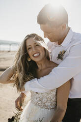 Affectionate bride and groom hugging on the beach at sunset - LHPF00792