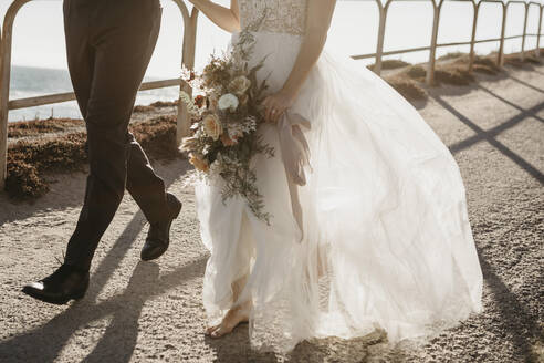 Close-up of bride and groom walking on path at the coast - LHPF00794