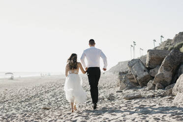 Rückansicht von Braut und Bräutigam beim Laufen am Strand - LHPF00804
