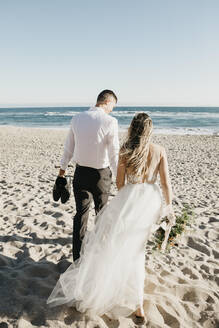 Rückansicht von Braut und Bräutigam beim Spaziergang am Strand - LHPF00808
