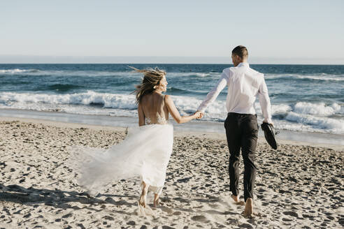 Rückansicht von Braut und Bräutigam beim Laufen am Strand - LHPF00810