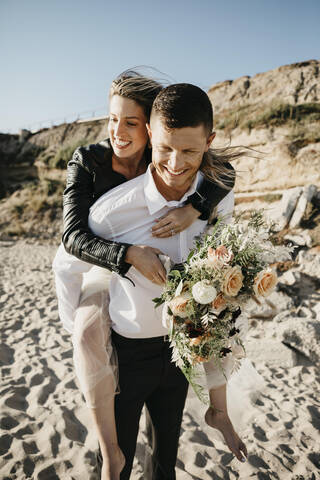 Glücklicher Bräutigam trägt Braut huckepack am Strand, lizenzfreies Stockfoto