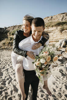 Glücklicher Bräutigam trägt Braut huckepack am Strand - LHPF00814