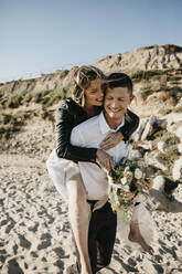 Happy groom carrying bride piggyback on the beach - LHPF00815