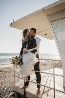Glückliche Braut und Bräutigam an der Rettungsschwimmerhütte am Strand bei Sonnenuntergang - LHPF00817