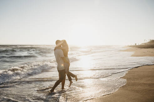 Zärtliches junges Paar, das sich bei Sonnenuntergang am Meeresufer umarmt - LHPF00834