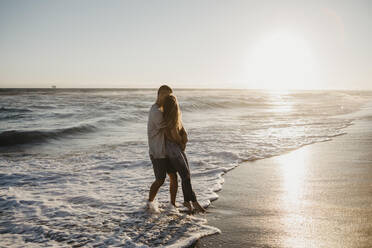 Zärtliches junges Paar, das sich bei Sonnenuntergang am Meeresufer umarmt - LHPF00835