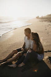 Glückliches junges Paar sitzt bei Sonnenuntergang am Strand - LHPF00837