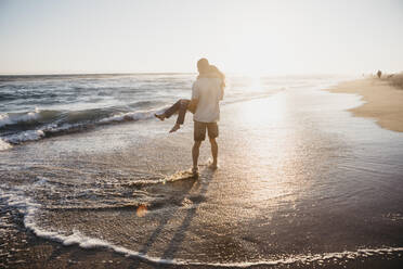 Happy young man carrying girlfriend at the seashore at sunset - LHPF00838