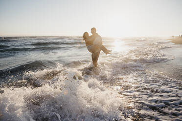 Happy young man carrying girlfriend at the seashore at sunset - LHPF00839
