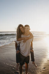 Portrait of happy young man carrying girlfriend piggyback on the beach at sunset - LHPF00840