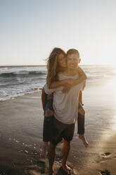 Portrait of happy young man carrying girlfriend piggyback on the beach at sunset - LHPF00841
