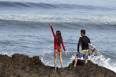 Rear view of a couple standing at the rocky beach, man with surfboard stock photo