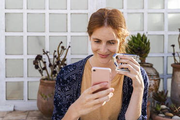 Portrait of smiling redheaded young woman with cup of coffee on terrace looking at cell phone - AFVF03953
