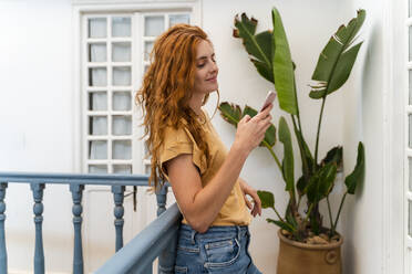 Smiling redheaded young woman leaning against railing looking at cell phone - AFVF03960