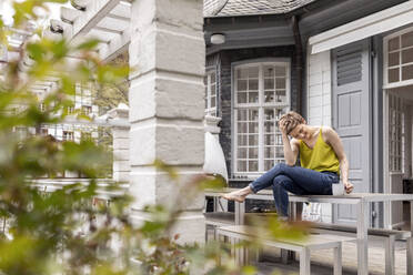 Entspannte Frau sitzt auf einem Terrassentisch im Garten ihres Hauses - MJFKF00005